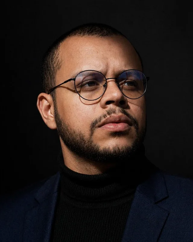 Michael Alves, con gafas redondas, jersey de cuello alto negro y americana azul marino